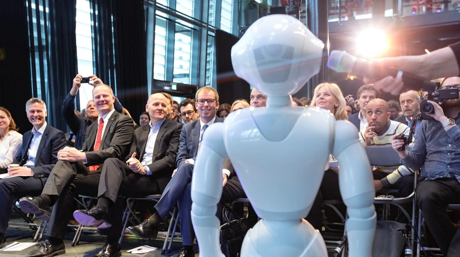 Bilde av en hvit robot som snakker i mikrofon foran sittende tilskuere.